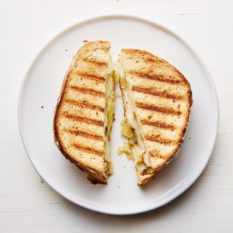 The Havarti & Caramelized Leek Grilled Cheese, sliced in half to reveal sweet, soft leeks and creamy melted Havarti, resting beside a bowl of red tomato soup.