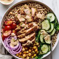 Roasted chicken and golden chickpeas top a bed of farro and fresh greens in a vibrant Lemon Tahini Chicken Power Bowl, drizzled with creamy sauce.  